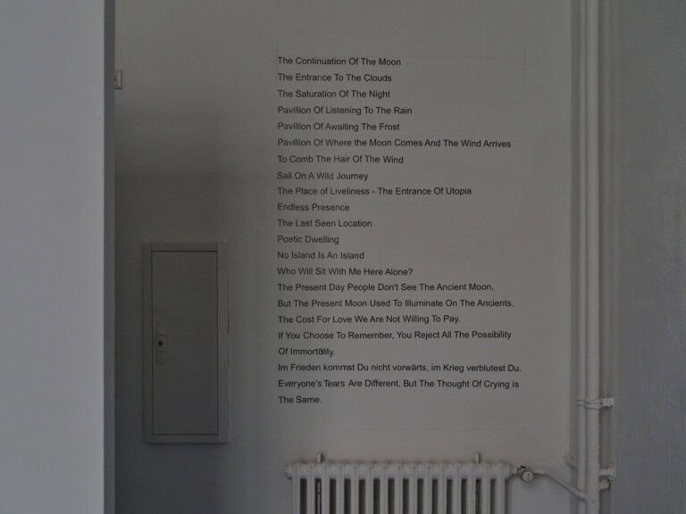 Foto: Ansicht eines Raumes mit weißer Wand. Auf der Wand befindet sich eine schwarzer Beklebung: "The Continuation Of The Moon/ The Entrance Of The Clouds/ The Saturation Of The Night/ Pavillion Of Listening To The Rain/ Pavillion Of Awaiting The Frosts/ Pavilion Of Where The Moon Comes And The Wind Arrives/ The Comb The Hair Of The Wind/ Sail On A Wild Journey/ The Place of Liveliness - The Entrance Of Utopia/ Endless Presence/ The Last Seen Location/ Poetic Dwelling/ No Island Is An Island/ Who Will Sit With Me Here Alone?/ The Present Day People Don't See The Ancient Moon,/ But The Present Moon Used To Illuminate On The Ancients./ The Cost For Love We Are Not Willing To Pay./ If You Choose To Remember, You Reject All The Possibility/ Of Immortality./ Im Frieden kommst du nicht vorwärts, im Krieg verblutest Du./ Everyone's Tears Are Different, But The Thought Of Crying is/ The Same.".