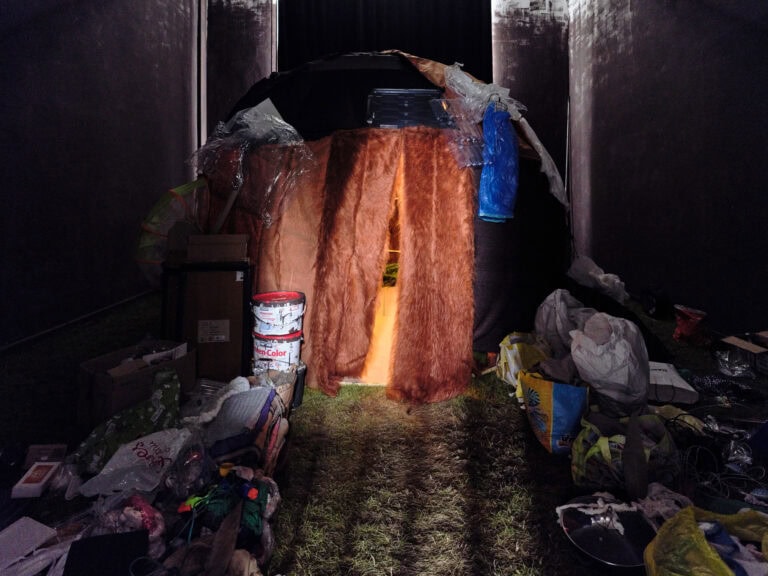 Foto: Ansicht eines Abgedunkelten Raumes. Der Boden des Raumes ist mit Rasen ausgelegt. Im Raum befindet sich eine große Menge an Dingen, die verteilt im Raum liegen. In der Mitte befindet sich ein Zelt, das aus unterschiedlichen Materialien besteht.