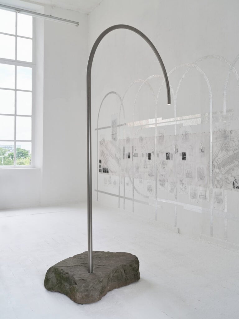 Foto: Nahaufnahme eines Steines auf weißem Boden vor einer weißen Wand. In den Stein ist eine Metallstange eingelassen. Die Wand ist mit Papieren und weiteren Materialien beklebt.