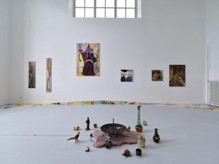 Foto: Ansicht eines Raumes mit weißen Wänden und grauem Boden. An den Wänden befinden sich mehrere Malereien im Klein-, Mittel- und Großformat. Ein Teil des Bodens, der Fußleiste und der Wand sind mit bunten Mustern bemalt. Auf dem Boden steht ein Arrangement aus mehreren Objekten.