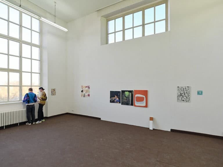 Foto: Ansicht eines Raumes mit braunem Boden, weißen Wänden und zwei Fenstern. An den Wänden befinden sich Malereien im klein- bis Mittelformat. Im Raum stehen zwei Menschen.