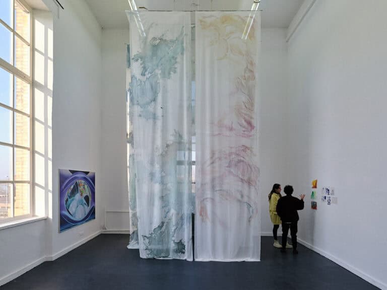 Foto: Ansicht eines Raumes mit dunklem Boden, weißen Wänden und einem großen Fenster. Im Raum Objekte, die an den Wänden hängen. Von der hohen Decke hängt ein Textil, das bis auf den Boden reicht. Im Raum befinden sich zwei Menschen.