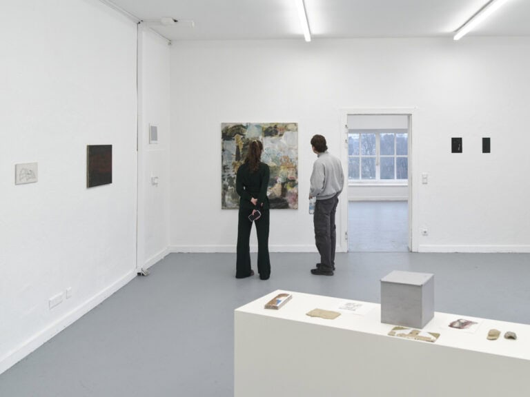 Foto: Ansicht eines Raumes mit weißen Wänden und grauem Boden. An den Wänden befinden sich Malereien in Klein- Mittel- und Großformaten. Im Vordergrund steht ein heller, länglicher Sockel, auf dem Objekte und Papiere liegen. Vor einer Leinwand stehen zwei Menschen, die die Arbeit in Nahsicht betrachten.