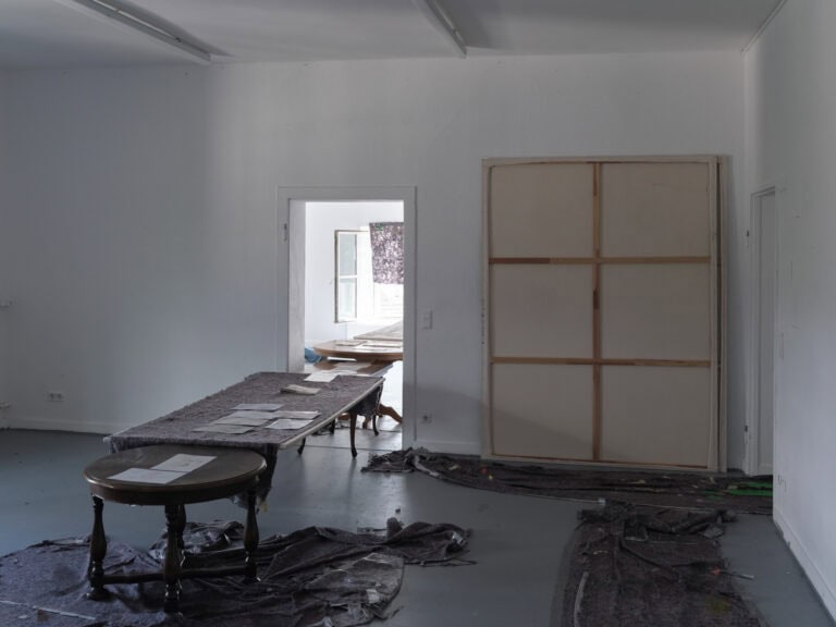 Foto: Ansicht eines Raumes mit weißen Wänden, grauem Boden und zwei Türen. Auf dem Boden liegt unregelmäßig verteilter und zusammengeschobener Malerfließ. An der Wand steht eine umgedrehte, großformatige Leinwand. Im Raum steht eine Reihe an Tischen, die durch die Tür hindurch in den nächsten Raum reicht. Einer der Tische ist ebenfalls mit Malerfließ bedeckt. Auf den Tischen liegen Papiere.