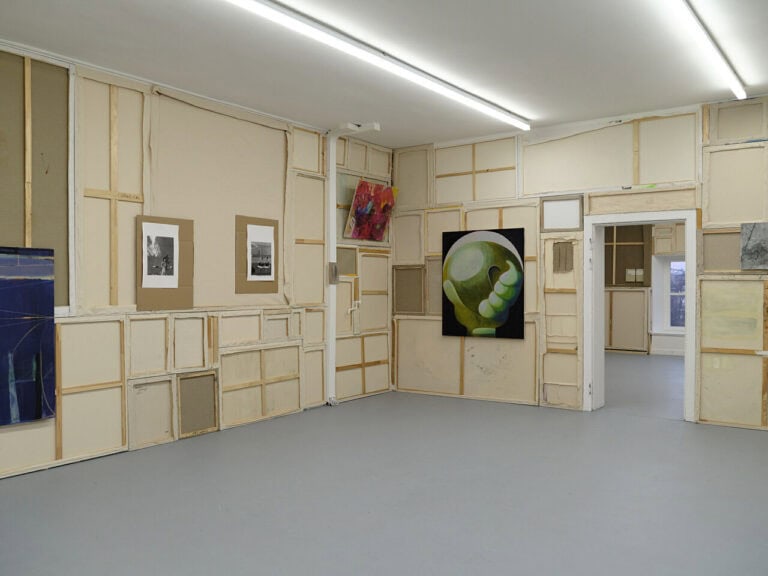 Foto: Ansicht eines Raumes mit grauem Boden. Die Wände sind mit umgedrehten Leinwänden vertäfelt. Auf den Leinwänden befinden sich weitere Objekte und Malereien.