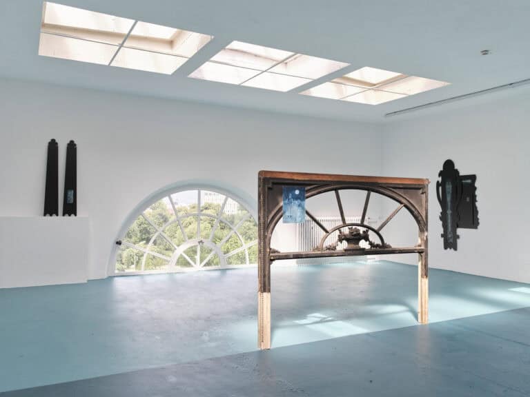 Foto: Ansicht eines Raumes mit weißen Wänden, einem kleinen Bogenfenster und Deckenfenstern. Der Boden ist in zwei unterschiedlichen Blautönen gestrichen. An den Wänden und im Raum befinden sich Objekte.