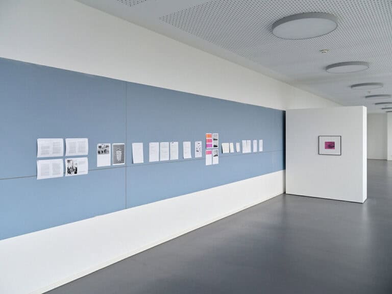 Foto: Raumansicht eines langen Raumes mit weißen Wänden und weißen Trennwänden. In der Mitte der Wand ein breiter, hellblauer Streifen. Darauf und auf den Stellwänden flache Objekte.