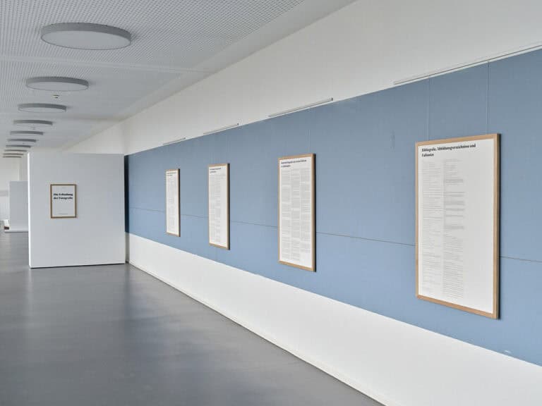 Foto: Raumansicht eines langen Raumes mit weißen Wänden und weißen Trennwänden. In der Mitte der Wand ein breiter, hellblauer Streifen. Darauf und auf den Stellwänden flache Objekte.