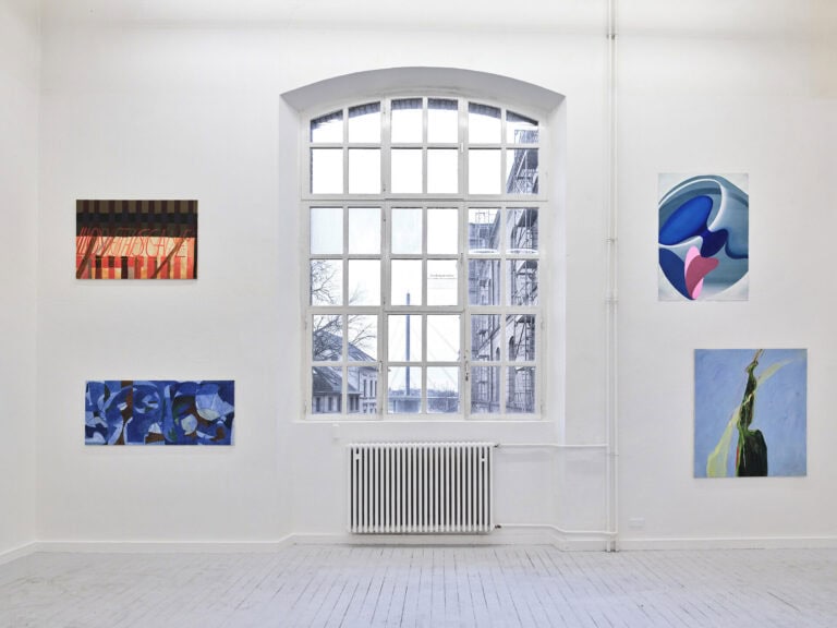Foto: Ansicht eines Raumes mit weißen Wänden, grauem Dielenboden und großem Bogenfenster. An den Wänden hängen, auf unterschiedlicher Höhe, Arbeiten auf Papier und Leinwand.