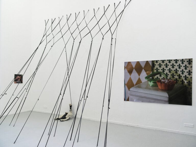 Foto: Ansicht eines Raumes mit weißen Wänden und grauem Boden. An den Wänden befinden sich zwei flache Objekte im Klein- und Großformat. Zwischen Wand und Boden sind mehrere miteinander verflochtene und geknotete schwarze Fäden gespannt. An einem der Fäden hält ein Objekt von der Decke, dass auf dem Boden liegt.