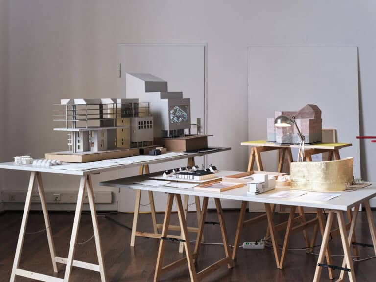 Foto: Ansicht eines Raumes mit weißen Wänden, einer Tür und Dielenboden. Ulm Raum stehen nah beieinander mehrere Tischplatten auf Böcken. Auf den Platten befinden sich architektonische Modelle, Lampen und Papiere.