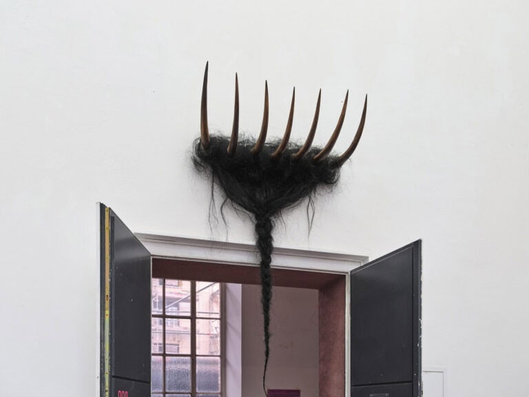 Foto: Ansicht eines Raumes mit weißer Wand und einer schwarzen, zweiflügligen, geöffneten Tür. Über der Tür hängt ein Objekt, dessen teile ihn den geöffneten Türrahmen hinein reichen.