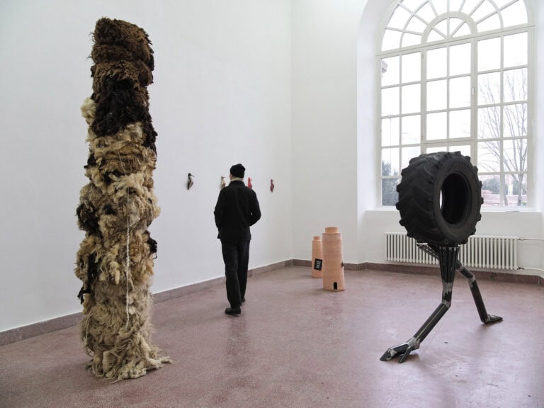 Foto: Ansicht eines Raumes mit weißem Wänden, gefliestem, rötlich-braunem Boden und Bogenfenster. Im Raum befinden sich drei große Skulpturen. An den Wänden hängen mehrere kleine Objekte. Durch den Raum bewegt sich ein Mensch.