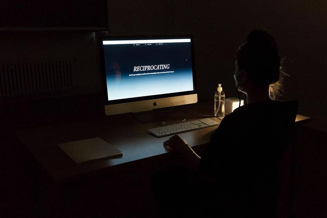 Foto: Ein dunkler Raum in dem ein Tisch steht. Auf dem Tisch steht ein eingeschalteter Computerbildschirm, auf dem Schrift zu sehen ist. Das Wort "RECIPROCATING" ist lesbar. Vor dem Tisch sitzt eine Person.