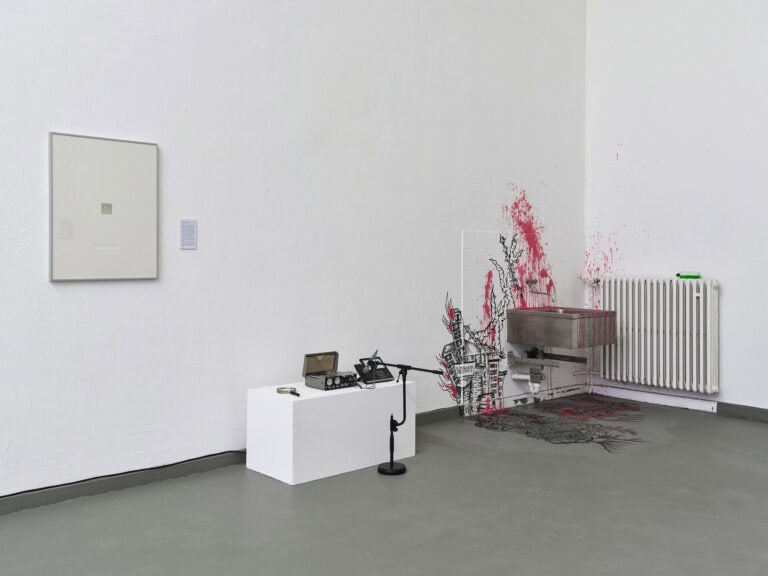 Foto: Ansicht eines Raumes mit weißen Wänden und grauem Boden. An der Wand befindet sich ein Objekt. Vor der Wand steht ein weißer Sockel, auf dem sich Objekte befinden und vor dem ein Mikrofon aufgebaut ist. In der Ecke des Raumes befindet sich ein Waschbecken und eine Heizung. Diese sind, von der Wand bis zum Boden, mit Farbsprenklern und Zeichnungen versehen.