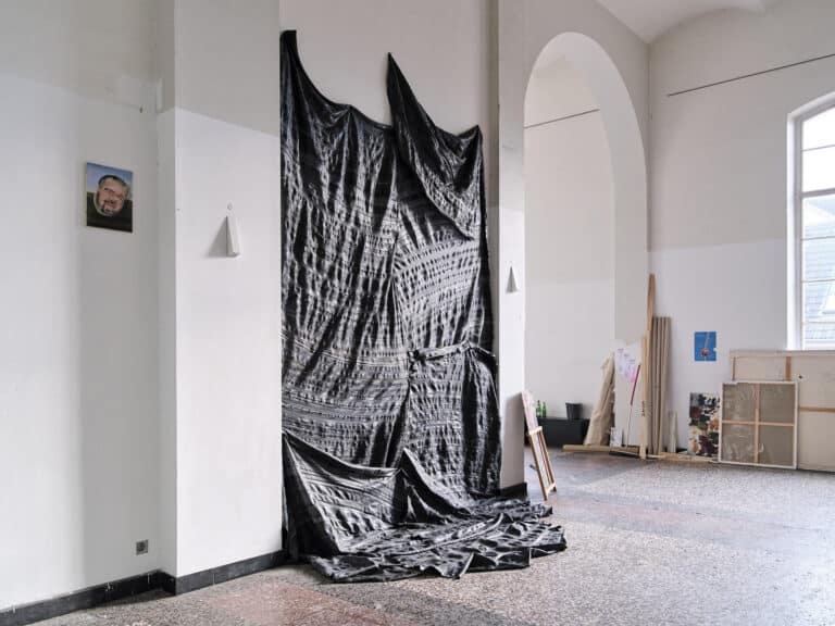 Foto: Ansicht eines Raumes mit weißen Wänden, schwarzer Fußleiste, mehrfarbige Steinboden und Fenster. An den Wänden hängen Malereien und faltenwerfendes Material. An die Wände gelehnt steht außerdem eine Vielzahl von Gegenständen.