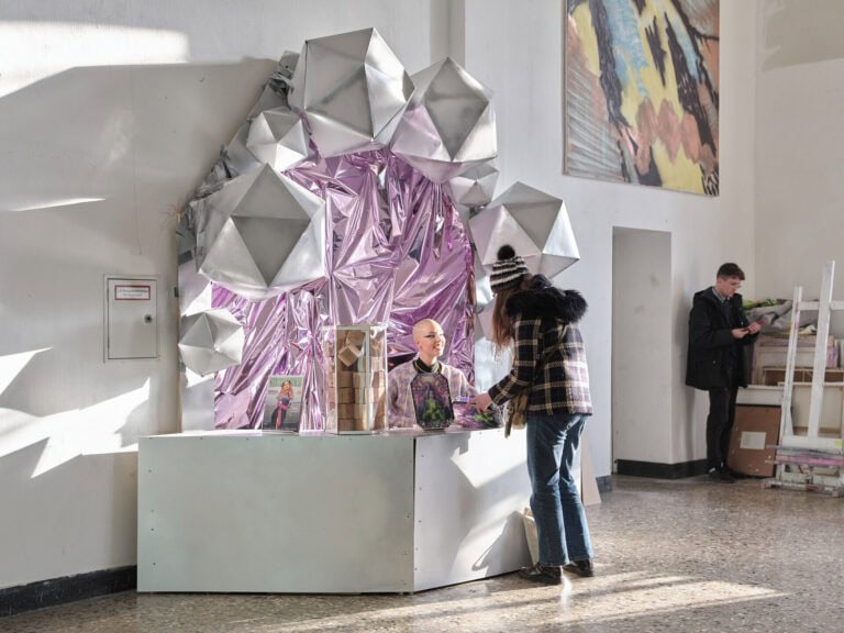 Foto: Ansicht eines Raumes mir weißen Wänden und mehrfarbigem Steinboden. Im Raum befindet sich eine Konstruktion aus silberfarbenem und lilalem, zum Teil glänzendem und reflektierendem Material. In der Konstruktion und im sonstigen Raum befinden sich insgesamt drei Menschen. An einer Wanden des Raumes befindet sich eine Malerei und an die Wände gelehnt weitere Objekte.