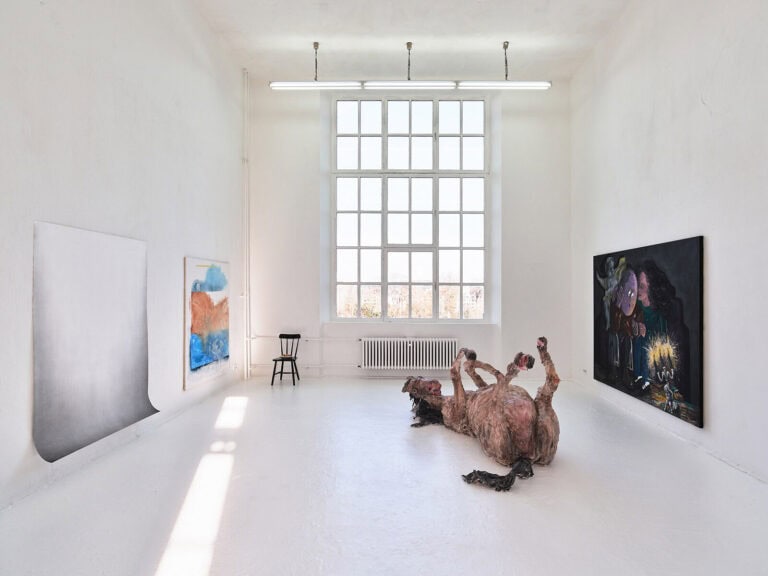 Foto: Sicht eines Raumes mit weißen Wänden, weißem Boden und einem großen Fenster. An den Wänden hängen drei großformatige Malereien und Papierarbeiten. Auf dem Boden liegt eine große Skulptur.