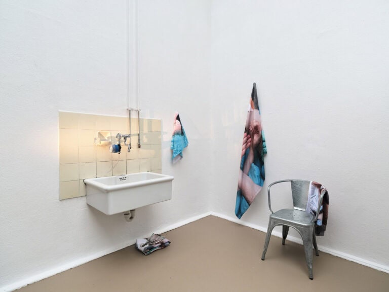 Foto: Ansicht eines Raumes mit weißen Wänden und beigem Boden. An der Wand befindet sich ein von Kacheln umgebenes Waschbecken und zwei bedruckte Textilien, die herunter hängen und Falten werfen. Unter dem Waschbecken befinden sich auf einem Stapel zusammengelegte, gefaltete Textilien. Vor der Wand steht ein Stuhl, auf dem ebenfalls eines der Textilien hängt. Die Fliesen überhalb des Waschbeckens werden von einem Lichtkegel angestrahlt.
