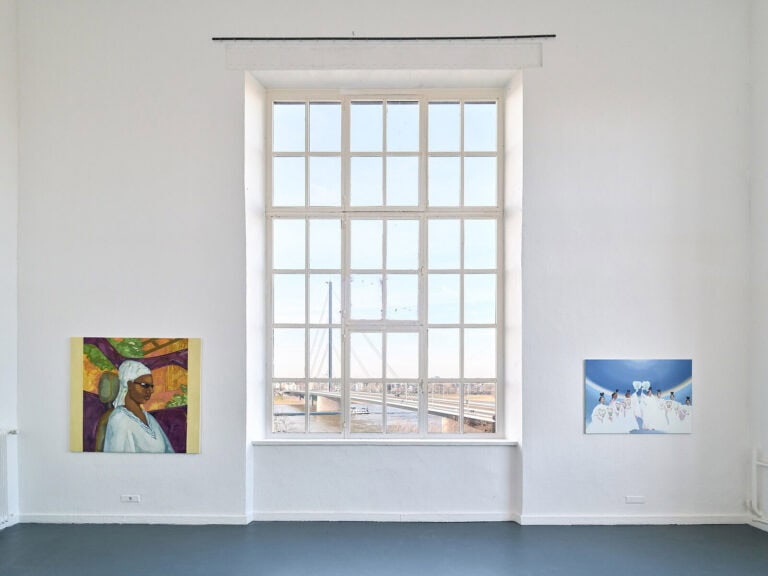 Foto: Ansicht eines Raumes mit weißen Wänden, grauem Boden und einem großen Fenster. An den Wänden hängen zwei mittelformatige Malereien. Durch das Fenster ist ein Fluss und eine Brücke zu sehen.