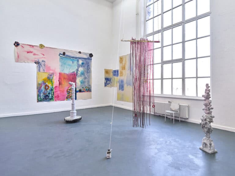Foto: Ansicht eines Raumes mit weißen Wänden, grauem Boden und einem großen Fenster. An den Wänden und auf dem Boden befinden sich Objekte. Ein Objekt hängt von der Decke und reicht bis kurz über den Boden.