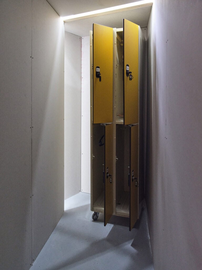 Foto: Ansicht eines schmalen, verschachtelten Raumes mit weißen Wänden, die aus Platten zusammengesetzt sind. Im Raum befindet sich ein hoher, schmaler Schrank auf Rollen mit vier geöffneten Türen.