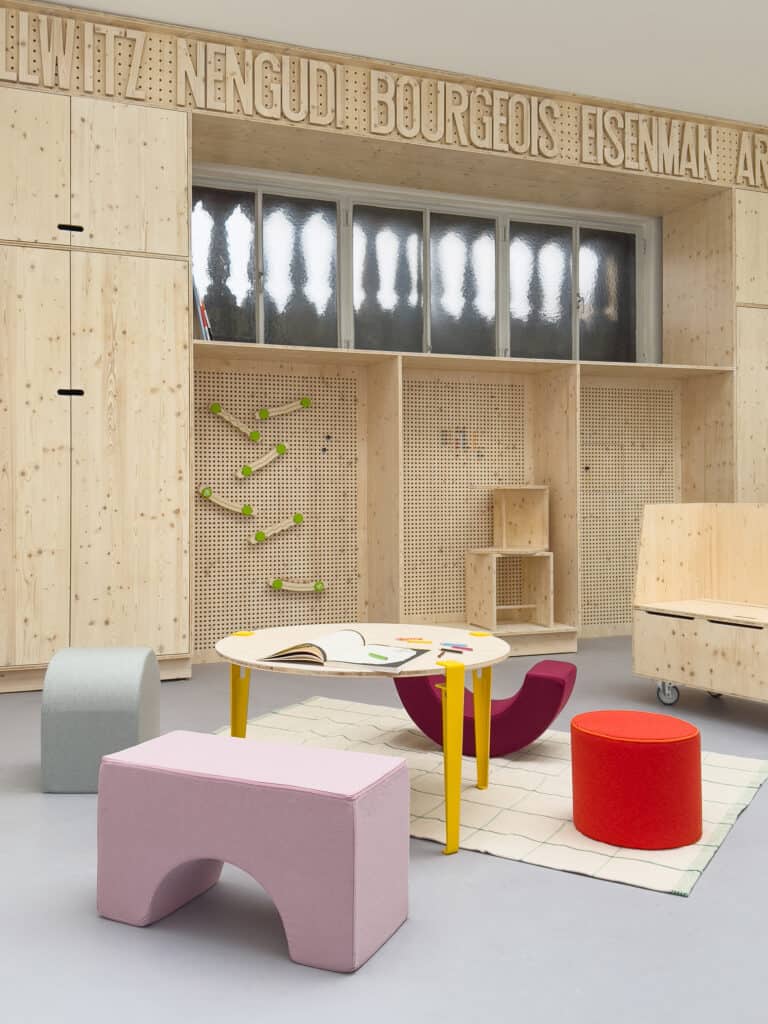 Foto: Raumansicht. Bunte Schemel um einen niedrigen Tisch, davor eine holzvertäfelte Wand mit Schriftzügen und einem Fenster.