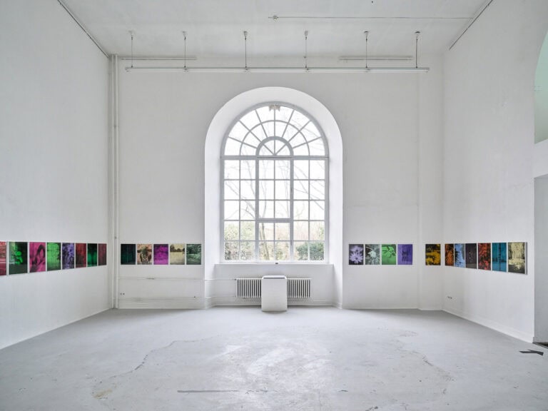 Foto: Ansicht eines Raumes mit weißen Wänden, grauem Boden und einem großen Bogenfenster. Im Raum hängen flache Objekte auf gleicher Höhe in einer durchgehenden Linie.