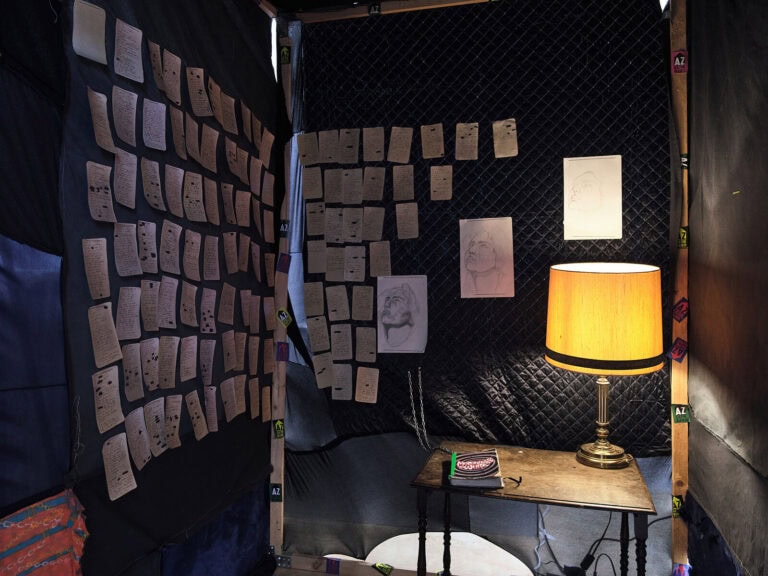 Foto: Ansicht einer Konstruktion in einem abgedunkelten Raum. An den eingezogenen Wänden hängen eine Vielzahl von Papieren mit Schrift und Zeichnungen. Auf einem kleinen Tisch steht eine eingeschaltete Lampe.