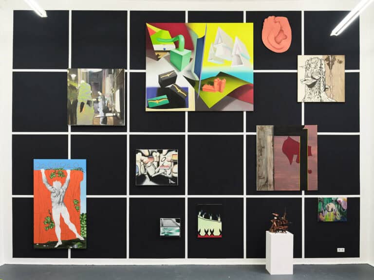 Foto: Ansicht eines Raumes mit grauem Boden. Die Wand ist mit schwarzen Quadraten in gleicher Größe versehen, die sich in gleichen Abständen zueinander befinden. Auf der Wand befinden sich, über- und Nebeneinander, Malereien in Klein-, Mittel- und Großformaten. Vor dem Wand steht ein weißer Sockel mit einem Objekt. gelblicher Wand und grauem Boden. An der Wand hängen Malereien im Klein-, Mittel- und Großformat, neben- und übereinander, auf unterschiedlicher Höhe. Rechts an der Wand befindet sich ein Waschbecken.