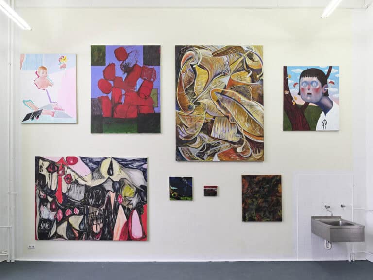 Foto: Ansicht eines Raumes mit gelblicher Wand und grauem Boden. An der Wand hängen Malereien im Klein-, Mittel- und Großformat, neben- und übereinander, auf unterschiedlicher Höhe. Rechts an der Wand befindet sich ein Waschbecken.