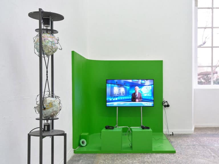 Foto: Ansicht eines Raumes mit weißen Wänden, mehrfarbigen Steinboden und Fenster. Im Raum befinden sich mehrere Objekte, darunter eine große Konstruktion in froschgrüner Farbe, in der sich ein Screen, Kopfhörer und Sitzgelegenheiten befinden.