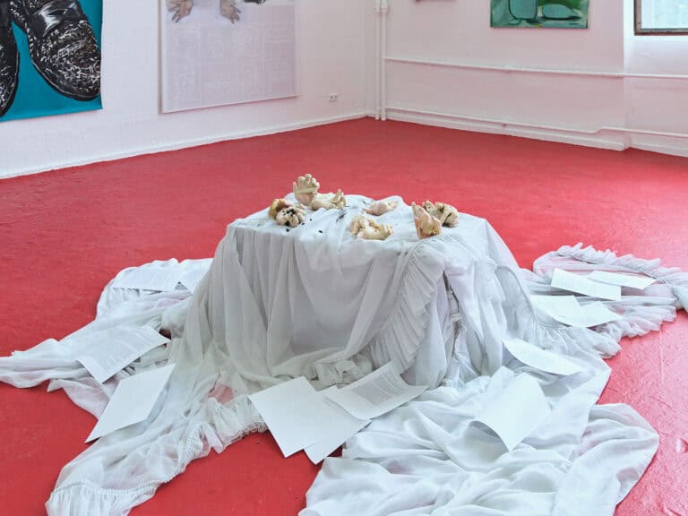 Foto: Nahaufnahme eines flächigen Objektes auf rotem Boden. Im Hintergrund in eine weiße Wand, auf der flache Objekte hängen.