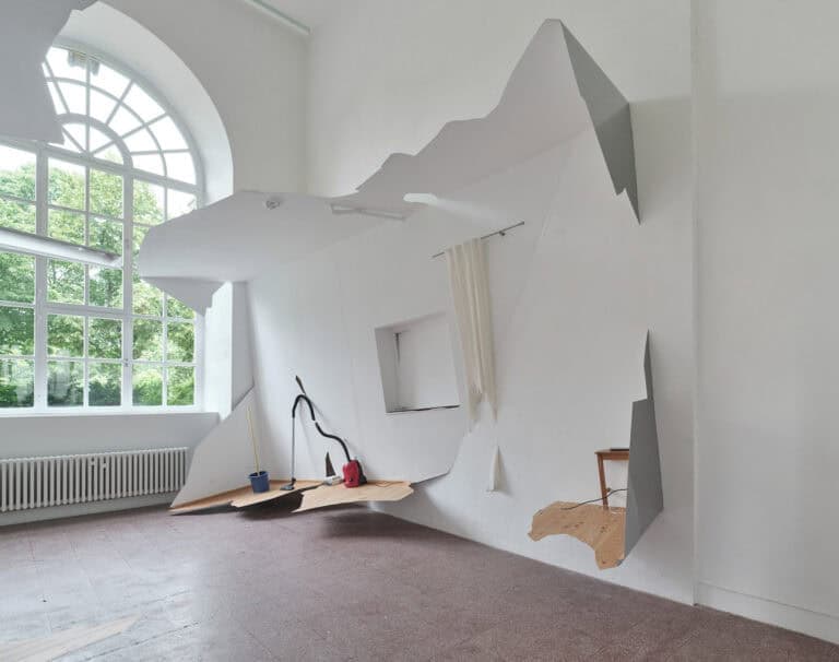 Foto: Ansicht eines Raumes mit weißen Wänden, einem roten, gekachelten Boden und einem großen Bogenfenster. An einer Wand ist eine Konstruktion aus unterschiedlichen Objekten angebracht, die sich in die Höhe über einen großen Teil der Wand erstreckt und die über dem Boden des Raumes schwebt.