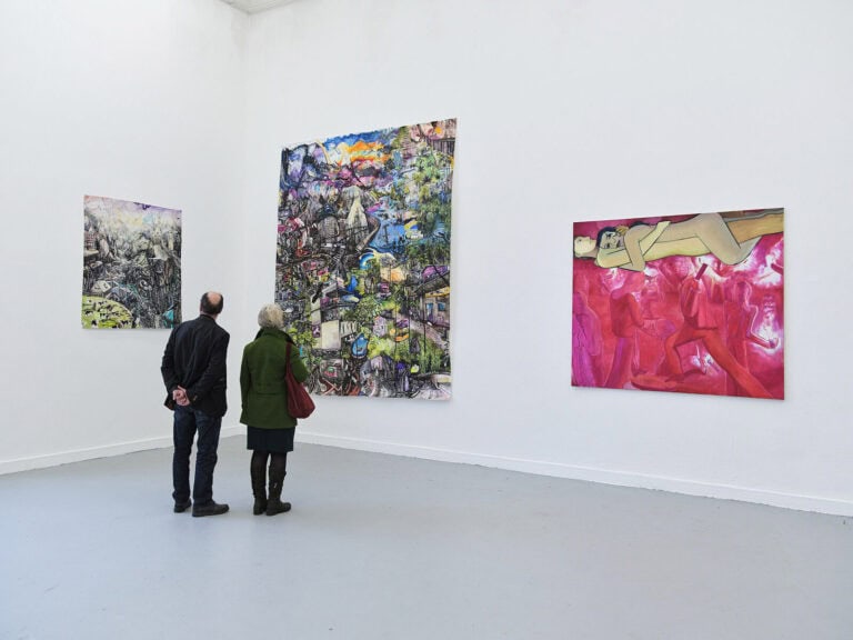 Foto: Ansicht eines Raumes mit weißen Wänden und grauem Boden. An der Wand hängen 3 Mittel- bis großformatige Leinwand und Papierarbeiten. Vor ihnen stehen zwei Menschen.