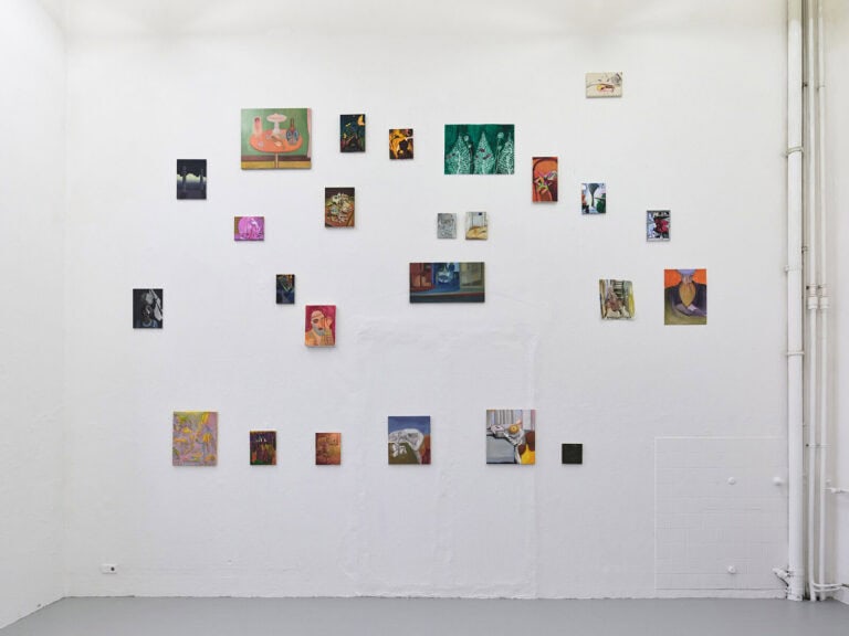 Foto: Ansicht eines Raumes mit weißen Wänden und grauem Boden. Über die Wand verteilt hängen, auf unterschiedlichen Höhen, in unterschiedlichen Abständen zueinander, bis unter die Decke, viele Malereien und Papierarbeiten in Klein- und Mittelformaten.